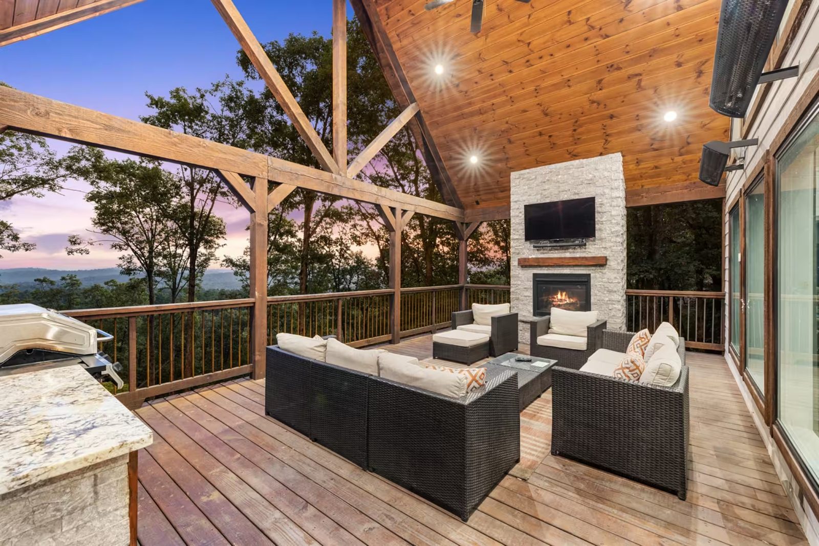Covered deck with a stone fireplace, lounge seating, and views over the treetops.